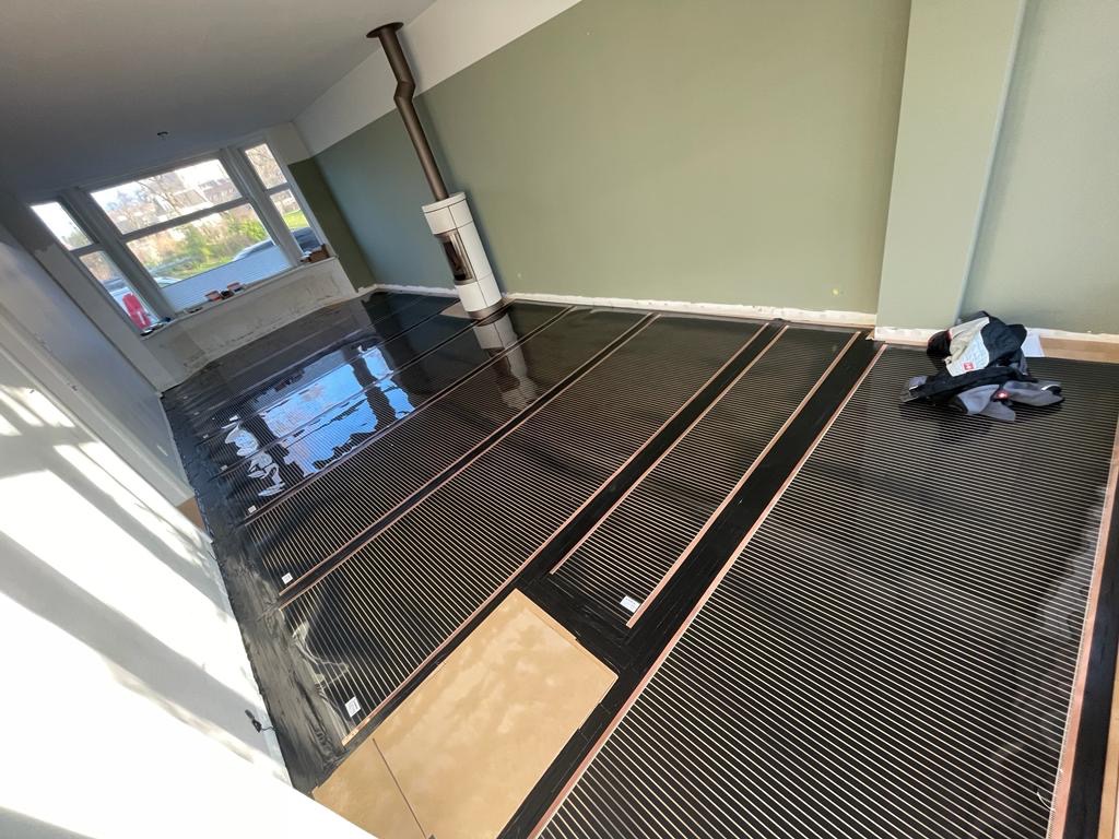 Rozłożone, czarne maty grzewcze z widocznymi miedzianymi przewodami na podłodze w pokoju z piecem wolnostojącym i oknem, przygotowane do dalszych prac wykończeniowych.