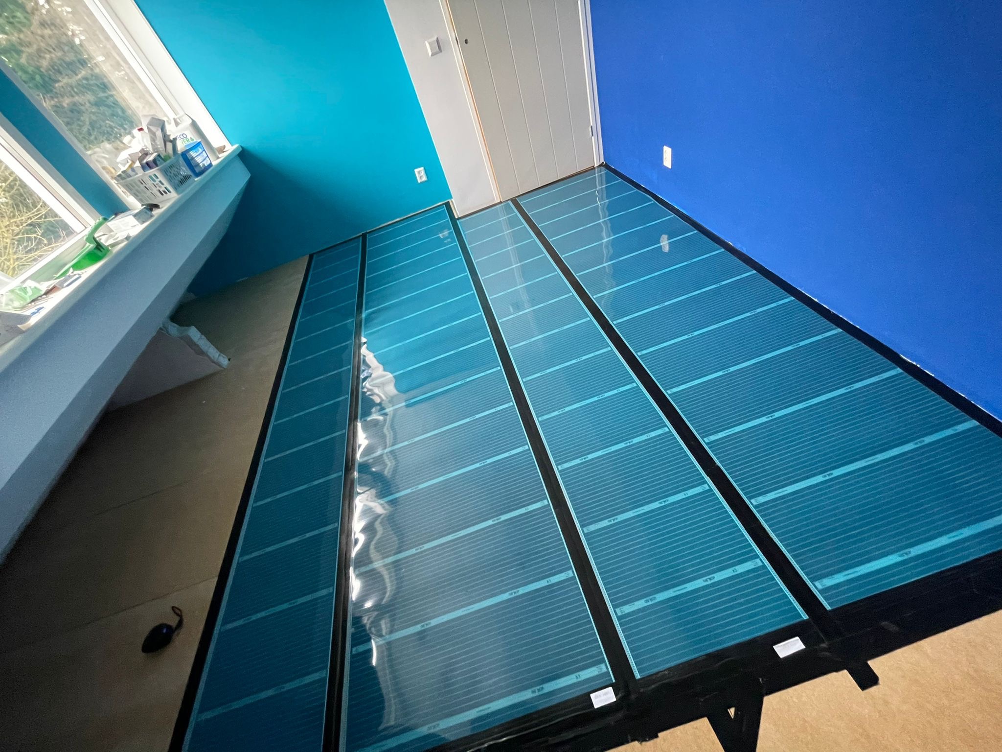 Niebieskie maty grzewcze rozłożone na podłodze w pomieszczeniu z niebieskimi ścianami, widok z dołu ukazuje strukturę mat i sposób ich ułożenia przed dalszymi pracami wykończeniowymi.