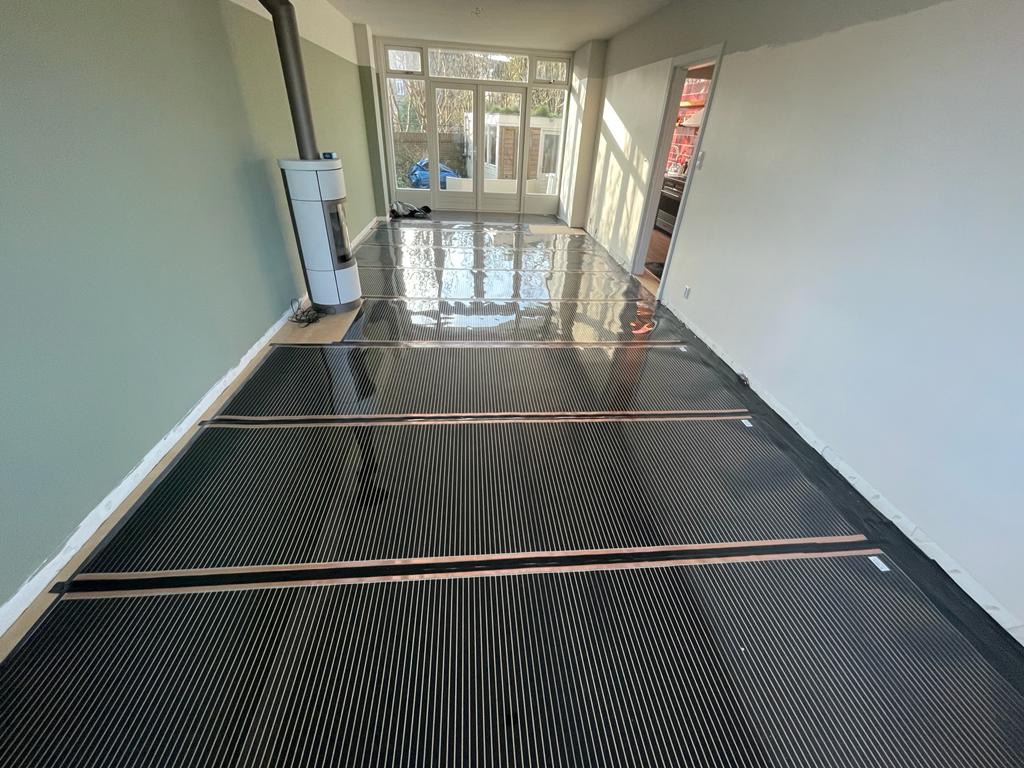 Rozłożone maty grzewcze na podłodze w jasnym pomieszczeniu z piecem wolnostojącym i dużymi oknami balkonowymi.