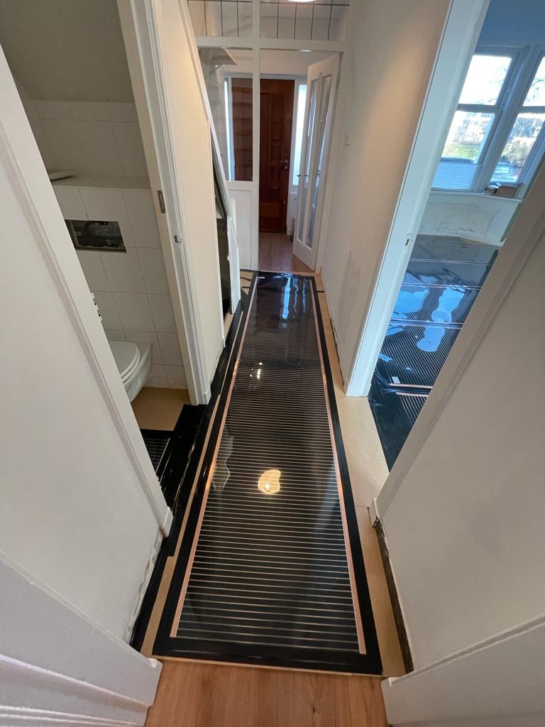 Widok z góry na czarną matę grzewczą z miedzianymi paskami, rozłożoną na drewnianej podłodze w wąskim korytarzu podczas instalacji, widoczne otwarte drzwi do łazienki i dalszej części domu.