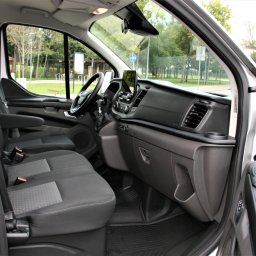 Seba-Trans Sebastian Myka - Wnętrze srebrnego busa z otwartymi drzwiami, widoczne czarne fotele z zagłówkami, deska rozdzielcza i fragment ulicy z zielenią za oknem.