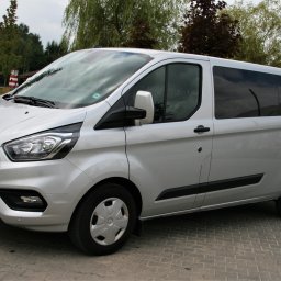Seba-Trans Sebastian Myka - Srebrny Ford Transit Custom Kombi z przyciemnianymi szybami, zaparkowany na brukowanej nawierzchni, z widoczną tablicą rejestracyjną i elementami zieleni w tle.