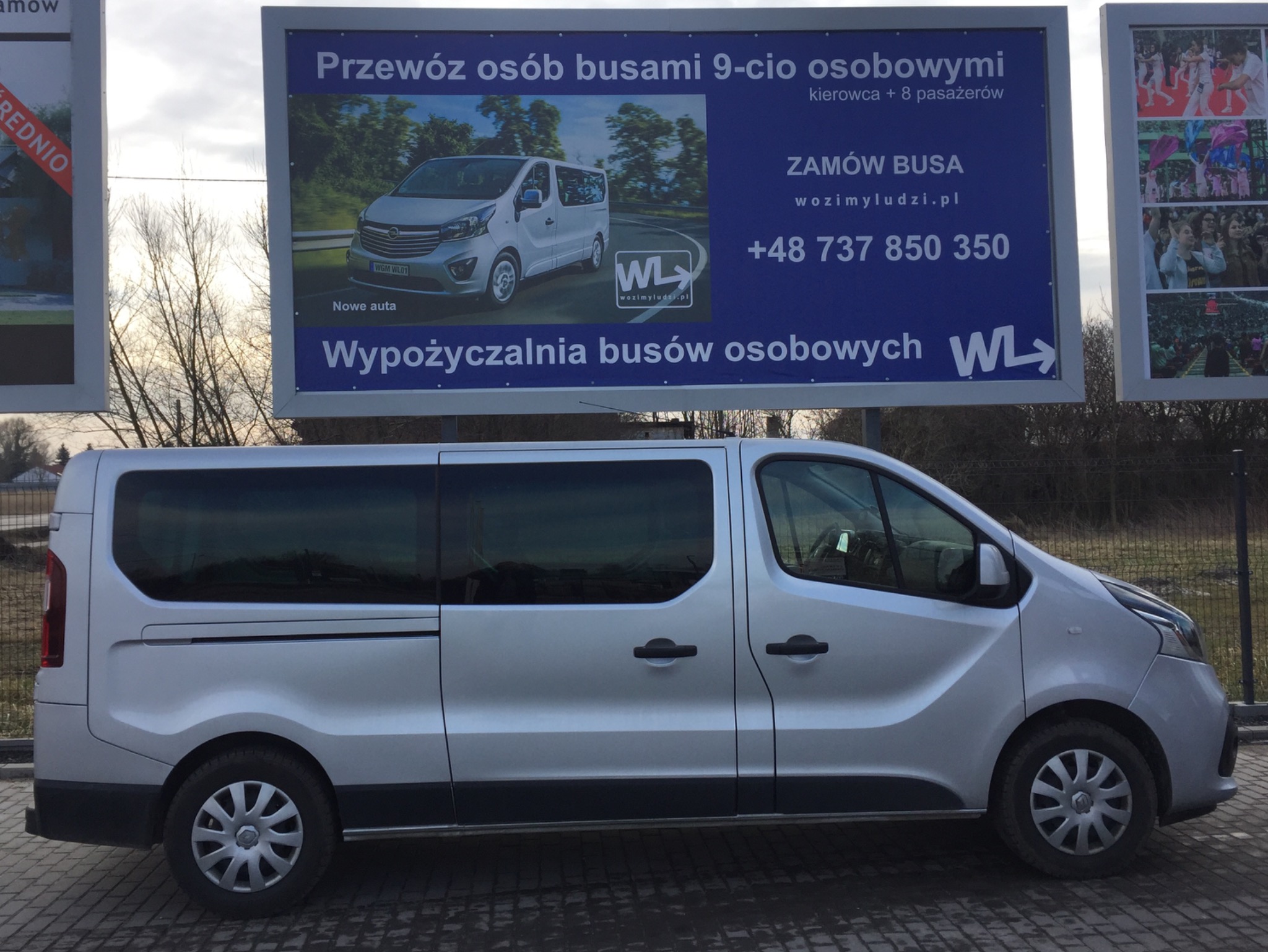 Srebrny bus osobowy zaparkowany przed billboardem reklamującym przewóz osób busami, z widocznym numerem telefonu i adresem strony internetowej.