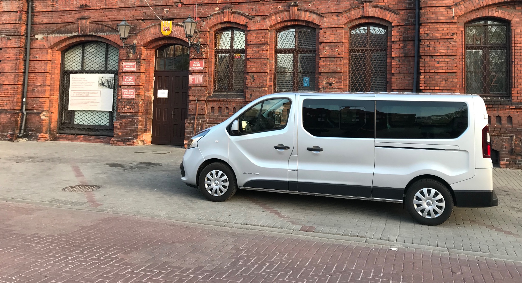 Srebrny bus transportowy zaparkowany przed budynkiem z czerwonej cegły w Żyrardowie, widoczny z boku, z refleksami światła na karoserii.
