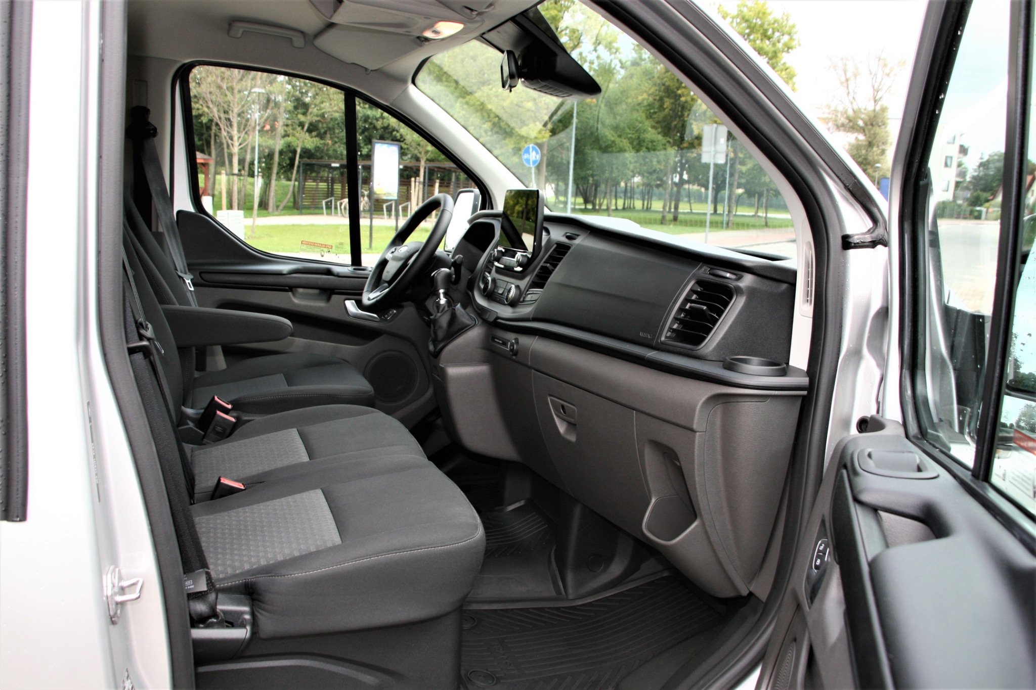 Wnętrze srebrnego busa z otwartymi drzwiami, widoczne czarne fotele z zagłówkami, deska rozdzielcza i fragment ulicy z zielenią za oknem.