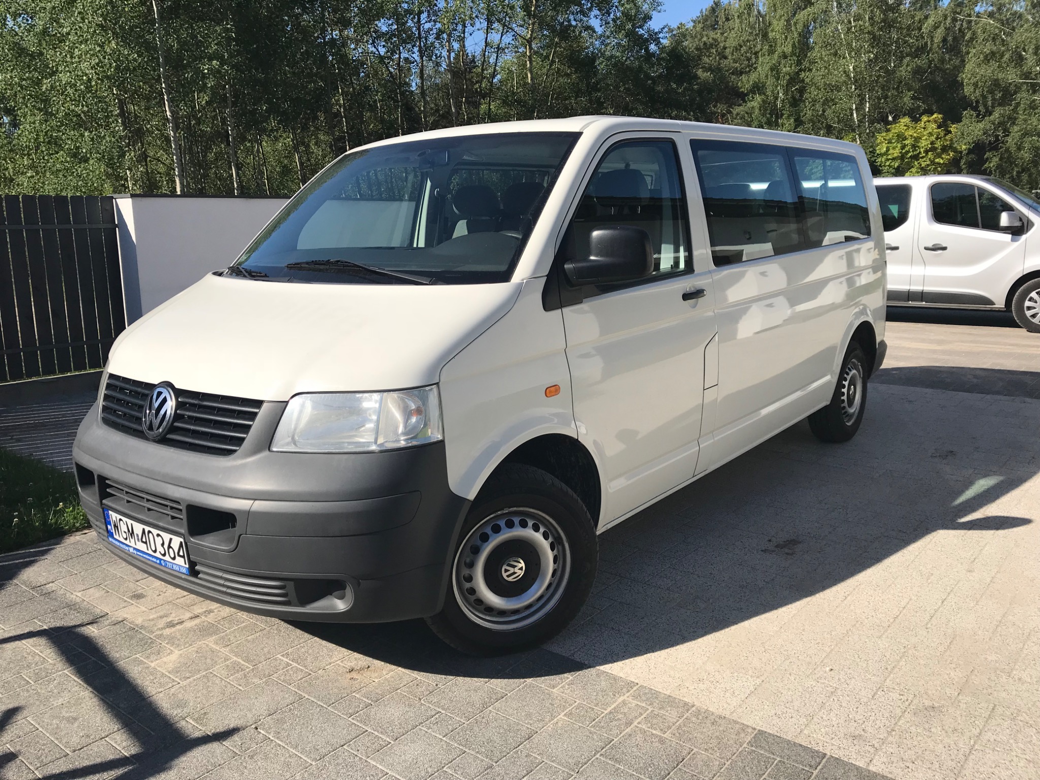 Biały Volkswagen Transporter T5 z ciemnoszarym zderzakiem, zaparkowany na kostce brukowej w słoneczny dzień, z widoczną tablicą rejestracyjną WGM 40364.