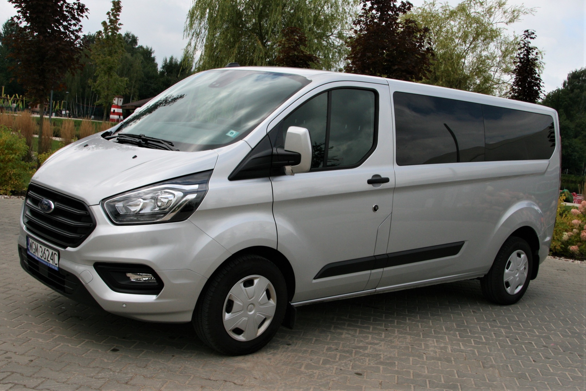 Srebrny Ford Transit Custom Kombi z przyciemnianymi szybami, zaparkowany na brukowanej nawierzchni, z widoczną tablicą rejestracyjną i elementami zieleni w tle.