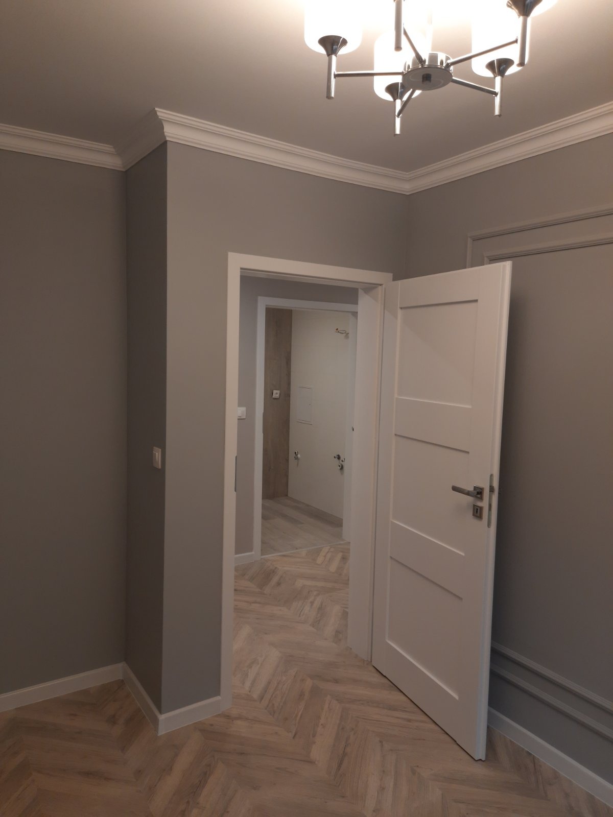 Wykończone wnętrze: szare ściany z białą sztukaterią, podłoga ułożona w jodełkę, białe drzwi wewnętrzne oraz widok na wykończoną łazienkę z jasnymi płytkami.