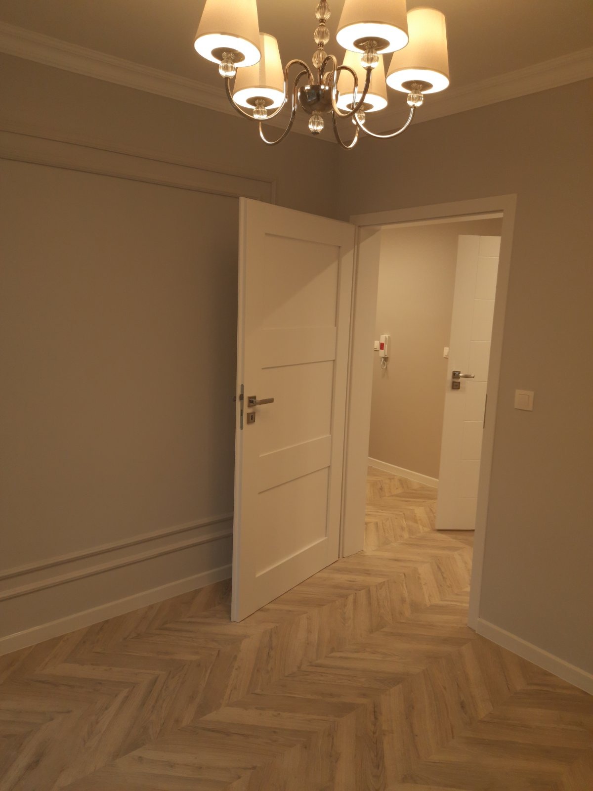 Nowoczesne wnętrze z parkietem w jodełkę, białymi drzwiami wewnętrznymi i ozdobną listwą przypodłogową, oświetlone wiszącym żyrandolem z abażurami.