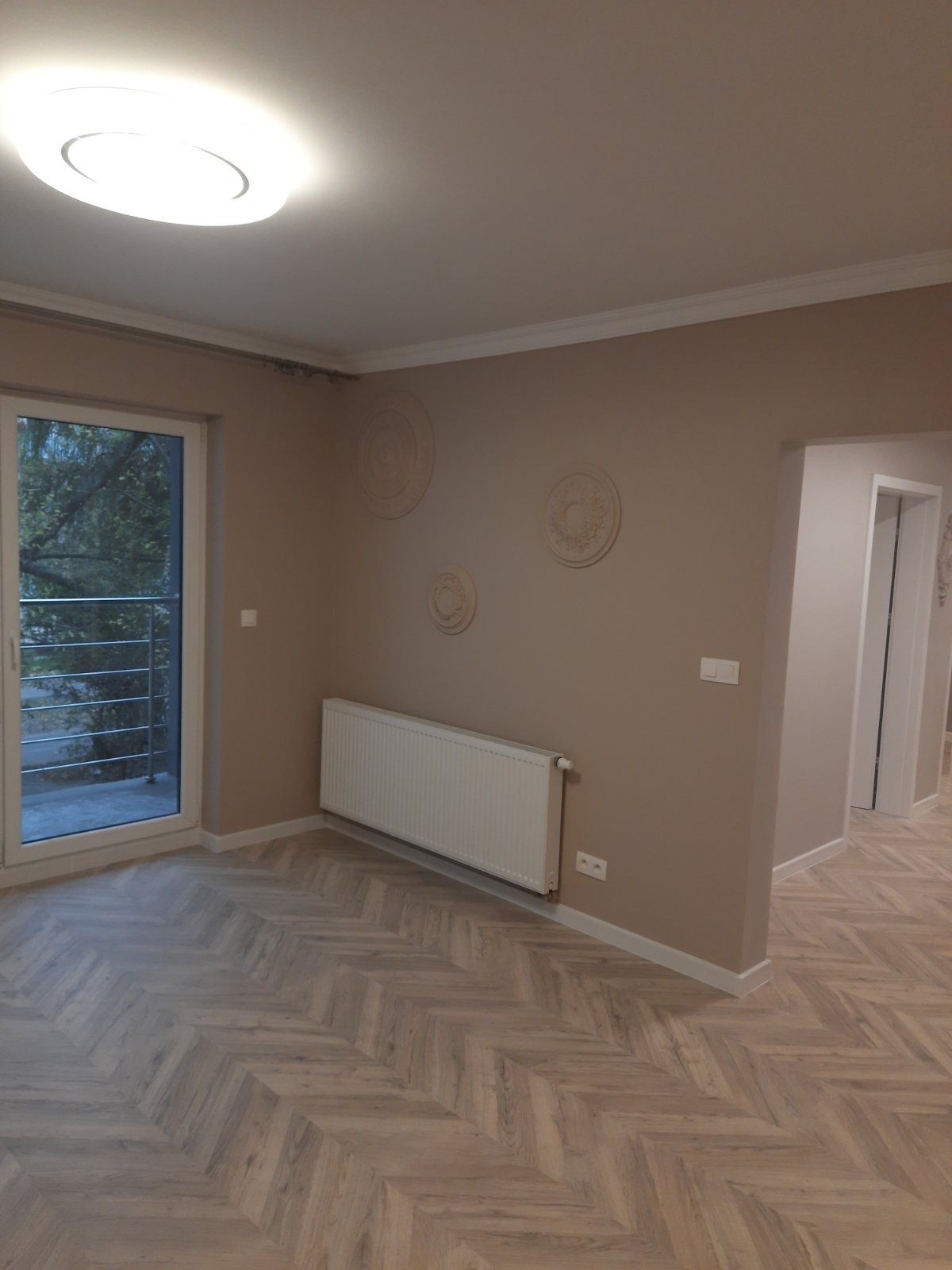 Pokój po remoncie z parkietem ułożonym we wzór jodełki, dekoracyjne rozety na ścianie, biały grzejnik i drzwi balkonowe.