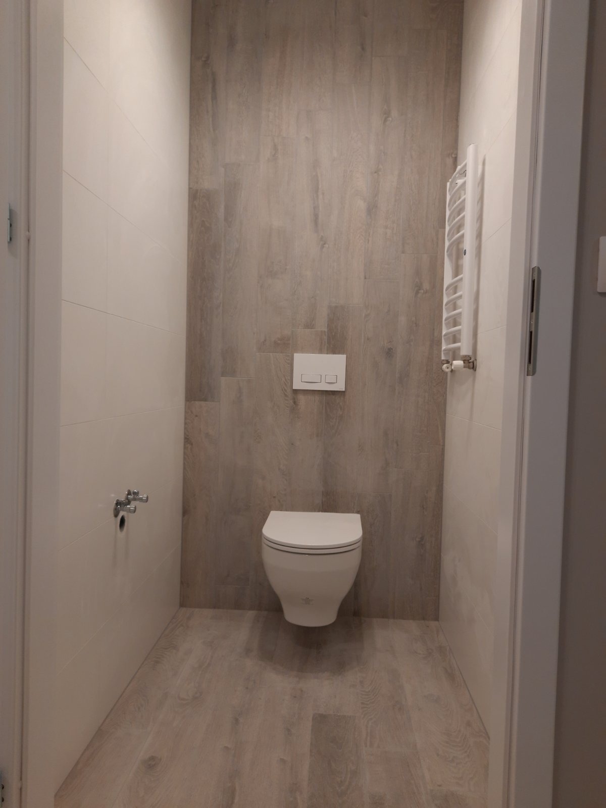 Nowoczesna łazienka z białymi płytkami na ścianach i panelami imitującymi drewno na podłodze i jednej ze ścian, wisząca toaleta i grzejnik drabinkowy.