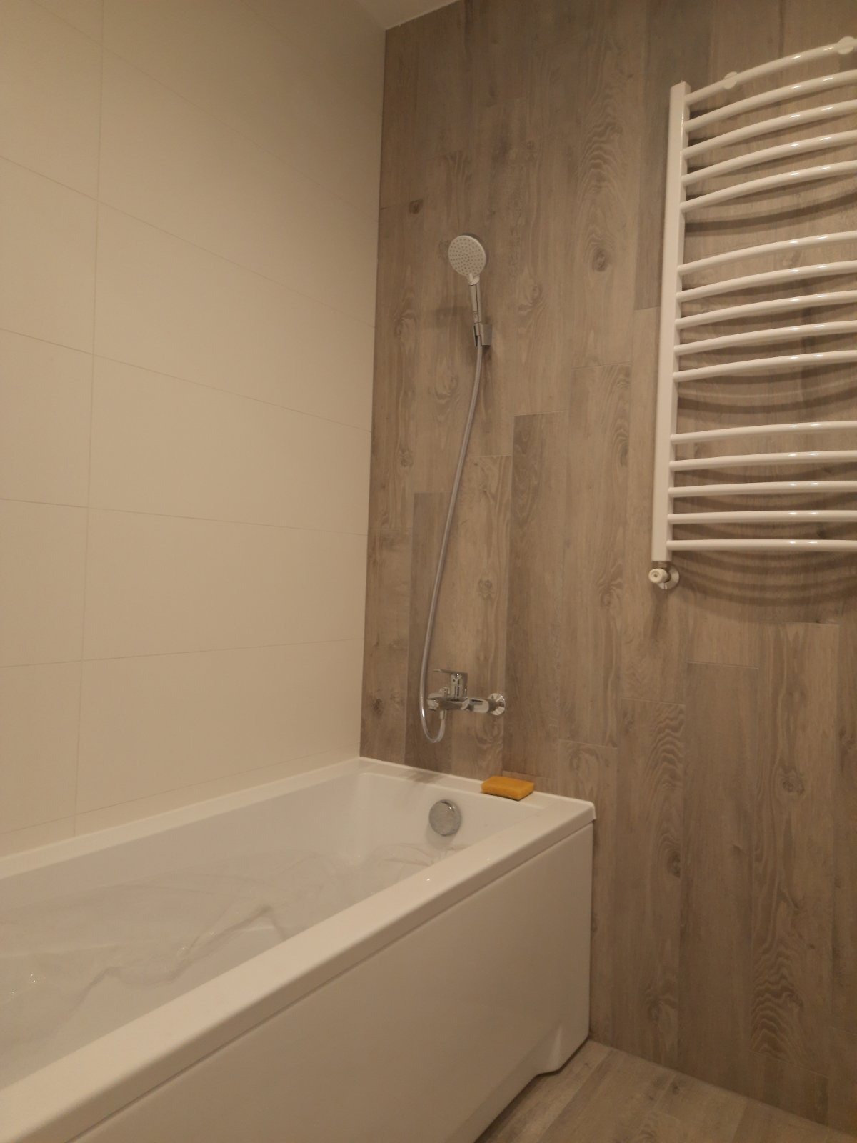 Biała wanna z chromowaną armaturą i słuchawką prysznicową na tle ściany wyłożonej panelami imitującymi drewno. Na brzegu wanny leży żółte mydło. Widoczny również biały grzejnik drabinkowy.