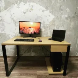 Biurko z blatem z jasnego drewna i czarnymi metalowymi nogami, z komputerem stacjonarnym, monitorem i laptopem, ustawione na tle ściany w szarym odcieniu betonu.