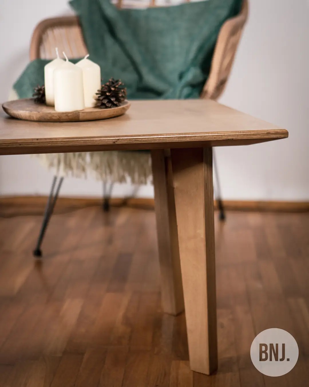 Stolik kawowy z jasnego drewna z trzema świecami i szyszkami na drewnianej tacy, w tle krzesło z poduszką i logo firmy w prawym dolnym rogu.