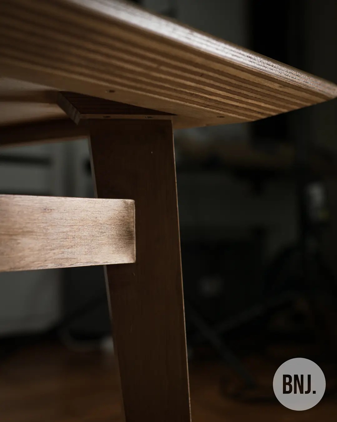 Detal drewnianego stołu z blatem z klejonego drewna warstwowego, widoczny fragment nogi i poprzeczki, ciemne tło.