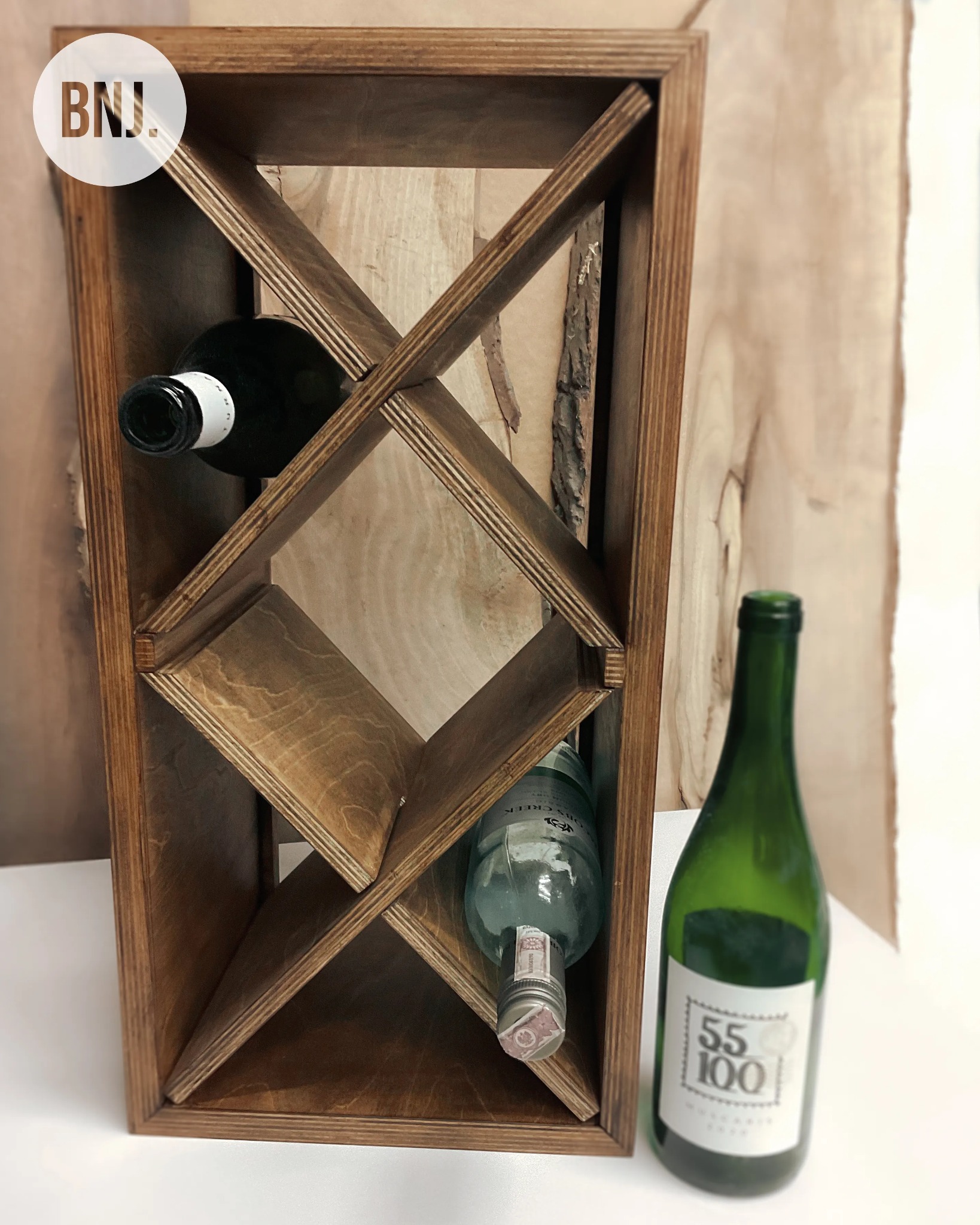 Drewniany regał na wino z butelkami, wykonany z jasnego drewna, z widocznymi warstwami sklejki na krawędziach, ustawiony na białym blacie na tle drewnianej płyty.