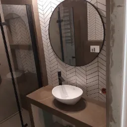 Nowoczesna łazienka z umywalką nablatową na brązowym blacie, okrągłe lustro z czarną ramą i białe płytki ułożone w jodełkę na ścianie, kabina prysznicowa z czarnymi profilami.