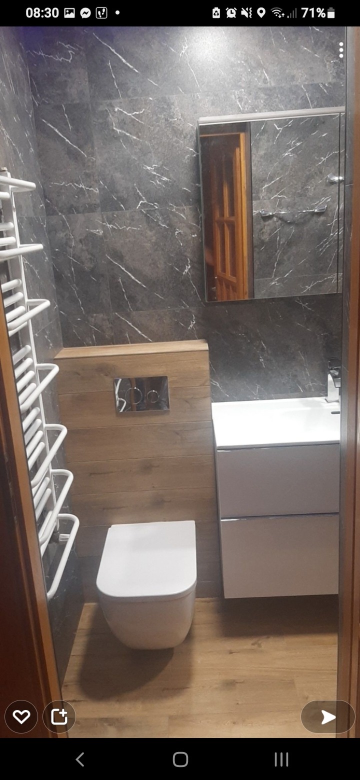 Nowoczesna łazienka z szarymi płytkami imitującymi marmur, drewnianymi panelami, podwieszaną toaletą, białą szafką z umywalką i grzejnikiem drabinkowym.