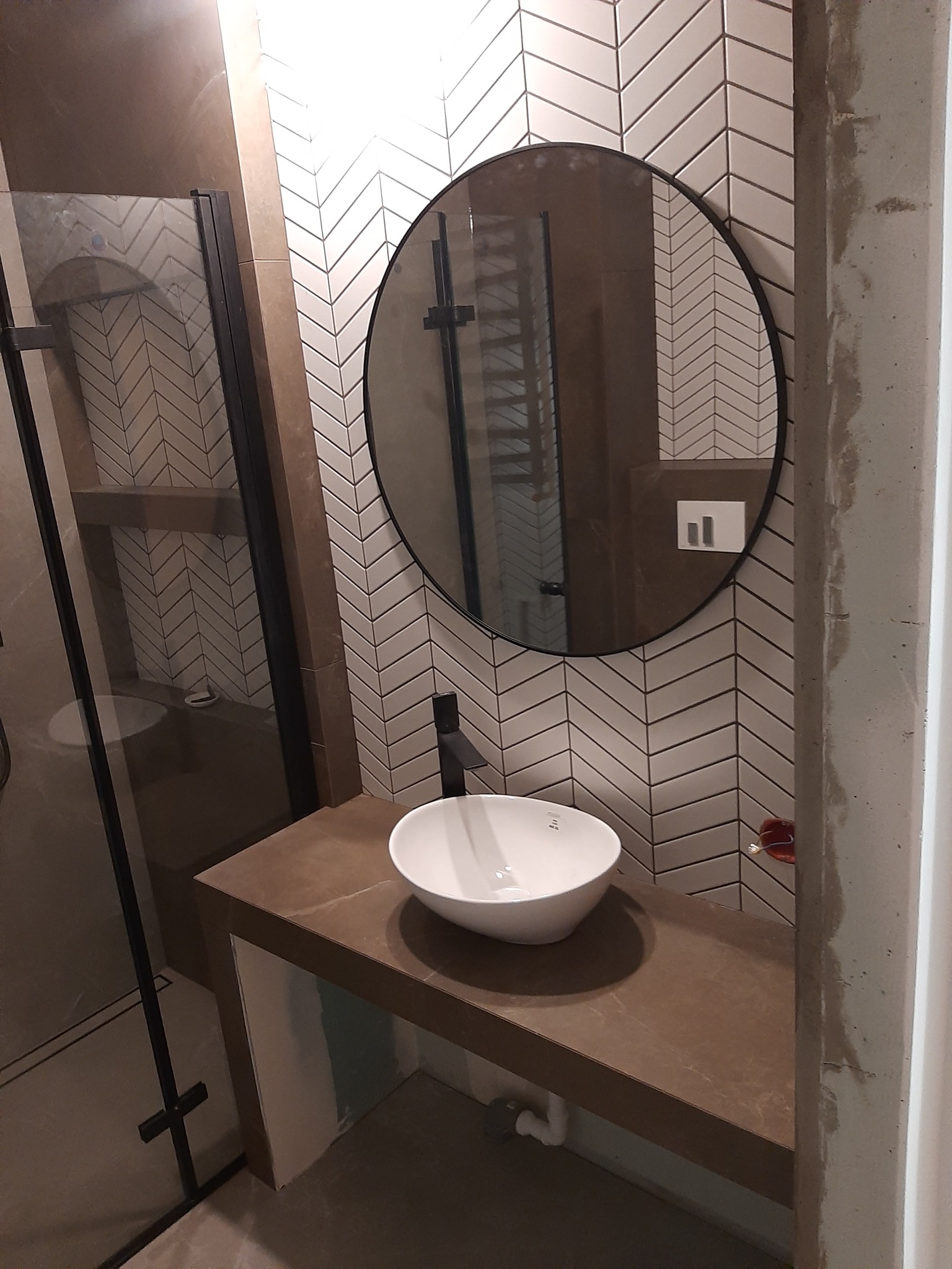 Nowoczesna łazienka z umywalką nablatową na brązowym blacie, okrągłe lustro z czarną ramą i białe płytki ułożone w jodełkę na ścianie, kabina prysznicowa z czarnymi profilami.