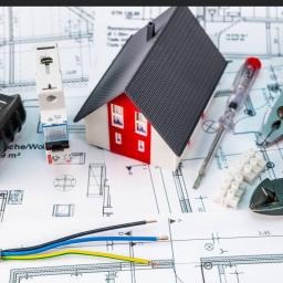 Makieta domu z czerwonymi ścianami na tle planów instalacji elektrycznej, obok bezpiecznik, przewody, zaciski, wkrętak i kombinerki.