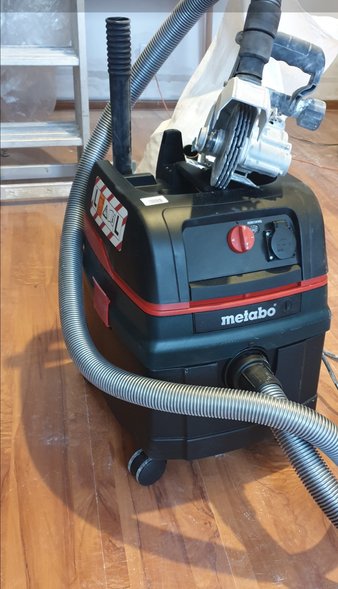 Odkurzacz przemysłowy Metabo z podłączoną bruzdownicą na drewnianej podłodze, widoczne ślady pyłu po cięciu.