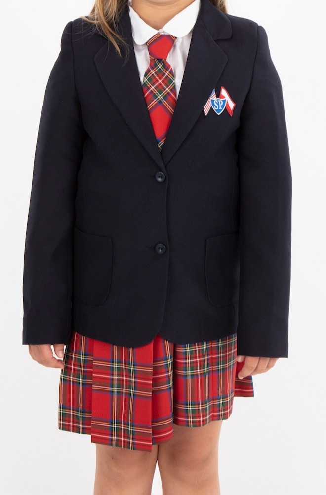 Dziewczynka w granatowej marynarce szkolnej, czerwonej spódnicy w kratę i pasującym krawacie, z przypinką z logo szkoły na klapie marynarki.