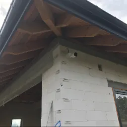 Biała kamera monitoringu zamontowana na rogu surowego budynku z białych bloczków, widoczne drewniane elementy dachu i czarna rynna