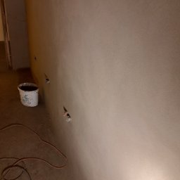 DR TYNK Dawid Radtke - Świeżo nałożony, gładki tynk na ścianie z widocznymi otworami na gniazdka elektryczne, wiadro po farbie i zwinięty kabel na podłodze.