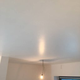 DR TYNK Dawid Radtke - Gładki, świeżo pomalowany sufit w jasnym odcieniu, oświetlony wiszącą żarówką bez klosza, z widocznymi ścianami w trakcie prac wykończeniowych i oknem zabezpieczonym folią.