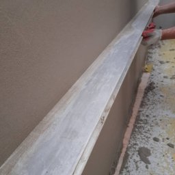 DR TYNK Dawid Radtke - Ręczne wyrównywanie świeżo nałożonego tynku na ścianie za pomocą długiej, aluminiowej łaty; widoczne czerwone rękawice robocze i fragment podłogi z resztkami zaprawy.