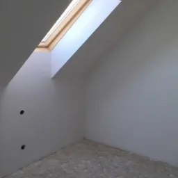 Poddasze z oknem dachowym w drewnianej ramie, ściany przygotowane do malowania, widoczne otwory na gniazdka elektryczne, podłoga zabezpieczona folią malarską.