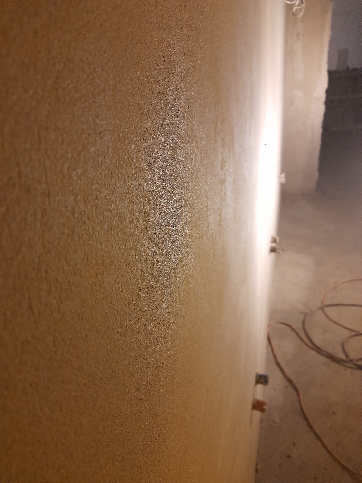 Świeżo nałożona, strukturalna warstwa tynku w odcieniach beżu, widoczna z bliska na ścianie w Rumi, z wystającymi puszkami elektrycznymi i zwisającym przewodem na podłodze.