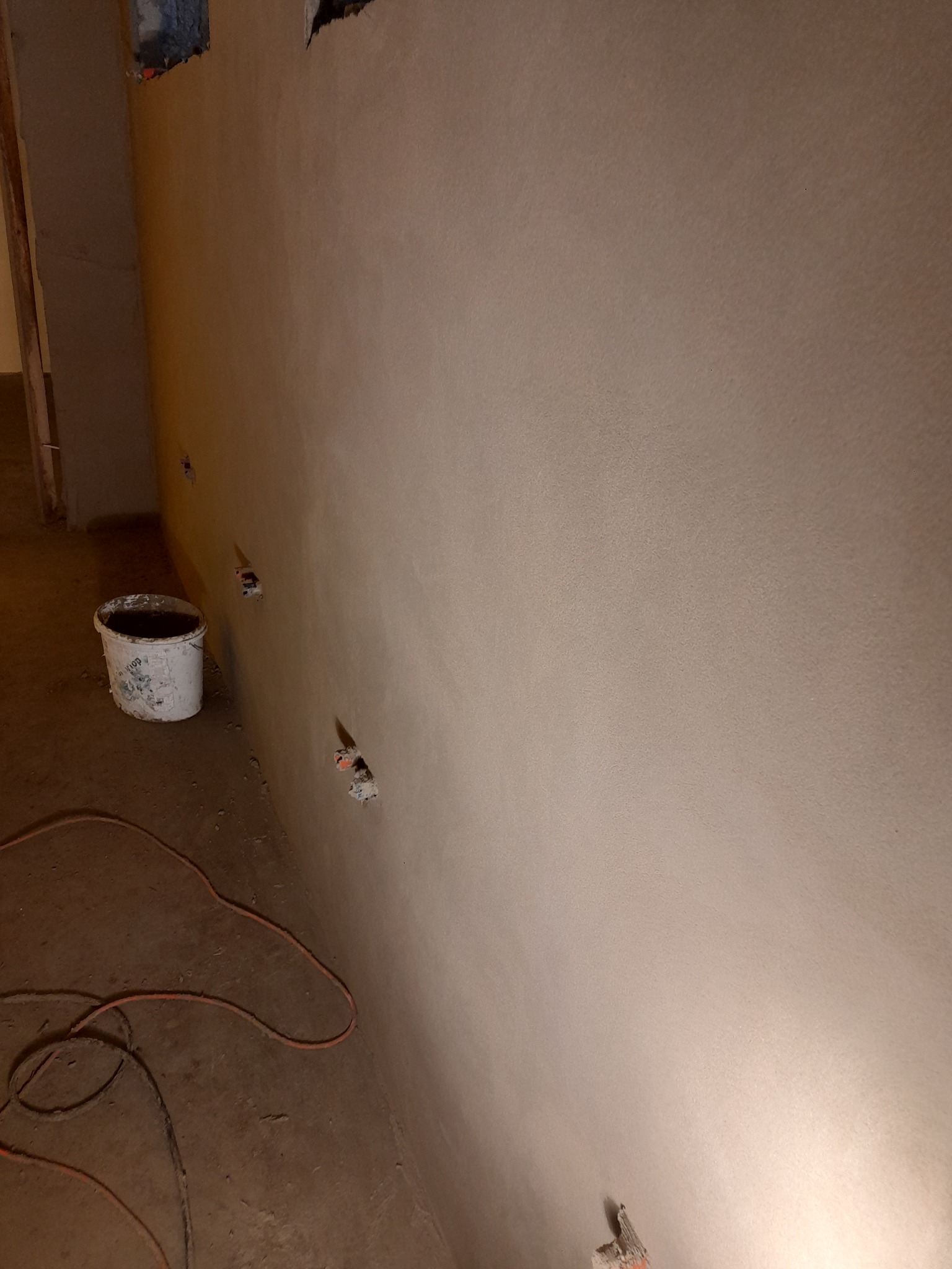 Świeżo nałożony, gładki tynk na ścianie z widocznymi otworami na gniazdka elektryczne, wiadro po farbie i zwinięty kabel na podłodze.