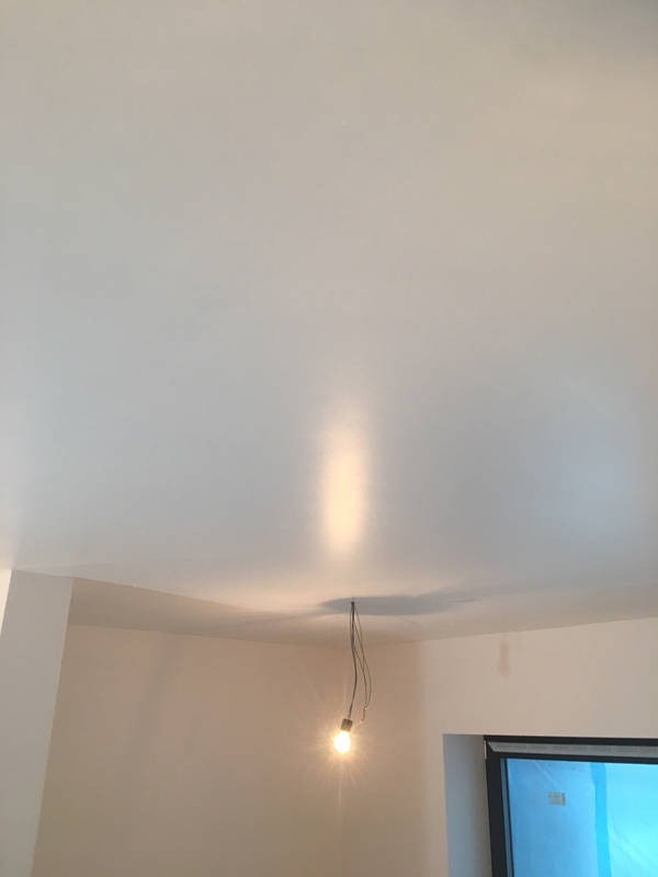 Gładki, świeżo pomalowany sufit w jasnym odcieniu, oświetlony wiszącą żarówką bez klosza, z widocznymi ścianami w trakcie prac wykończeniowych i oknem zabezpieczonym folią.