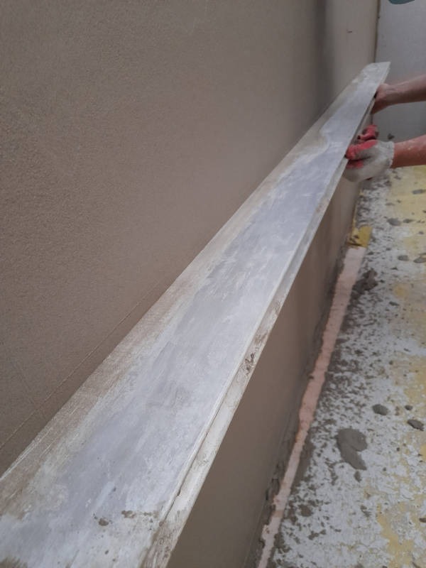 Ręczne wyrównywanie świeżo nałożonego tynku na ścianie za pomocą długiej, aluminiowej łaty; widoczne czerwone rękawice robocze i fragment podłogi z resztkami zaprawy.
