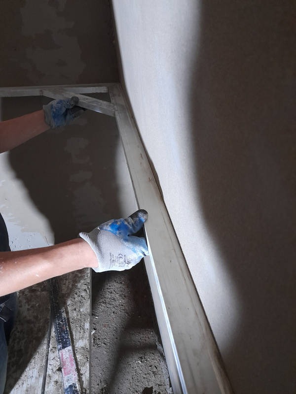 Ręka w rękawicy roboczej wygładza świeżo nałożony tynk na ścianie za pomocą długiej, aluminiowej łaty, widoczny fragment podłogi z resztkami zaprawy.