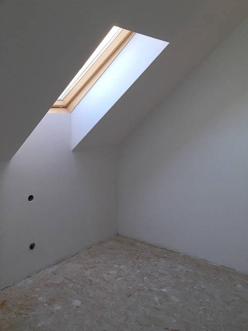 Poddasze z oknem dachowym w drewnianej ramie, ściany przygotowane do malowania, widoczne otwory na gniazdka elektryczne, podłoga zabezpieczona folią malarską.