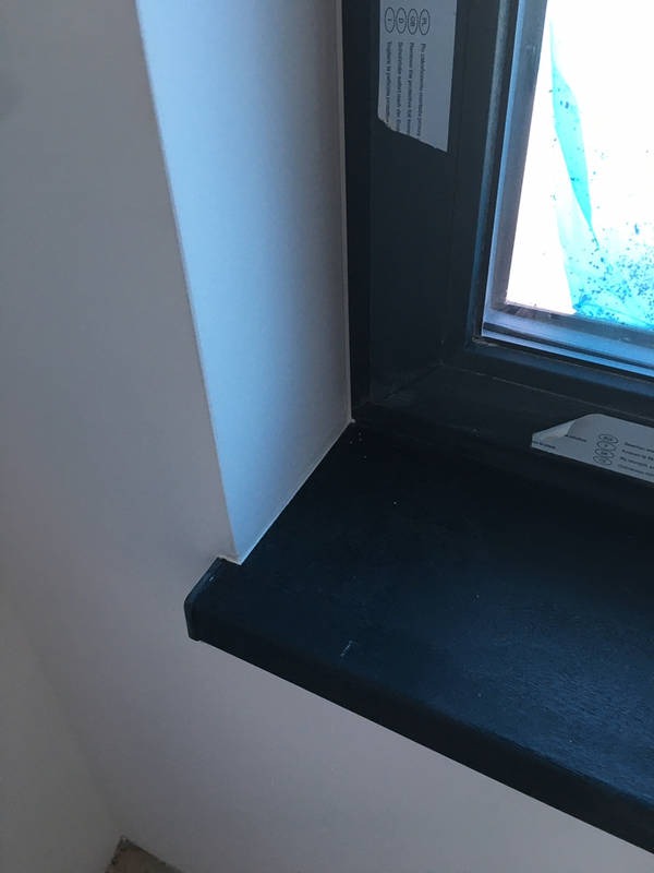 Fragment wnętrza z oknem o ciemnej ramie i białym parapetem, ściana pomalowana na biało, widoczne delikatne łączenie ściany z parapetem.