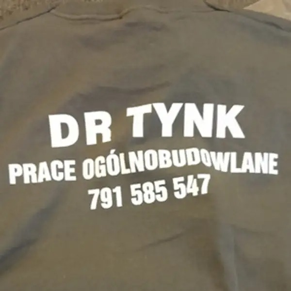 Fragment roboczej bluzy lub koszulki z nadrukowanym logo firmy 'DR TYNK', oferującej prace ogólnobudowlane, oraz numerem telefonu 791 585 547.