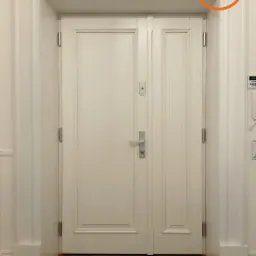 Białe, dwuskrzydłowe drzwi z ozdobną ramą i drewnianą podłogą, widoczny zamek i wizjer, u góry logo firmy.