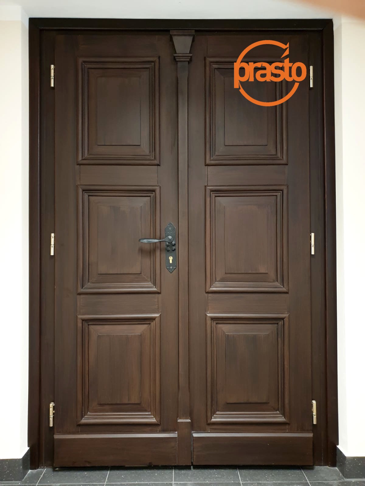 Dwuskrzydłowe, ciemnobrązowe drzwi z dekoracyjnymi panelami i czarną klamką, widoczne logo firmy na górze prawego skrzydła.