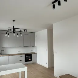 Nowoczesna, minimalistyczna kuchnia z szarymi szafkami, drewnianym blatem i podłogą, oświetlona industrialną lampą wiszącą i reflektorami sufitowymi, z białym stołem na pierwszym planie.