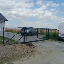 Nowo zamontowana, przesuwna brama wjazdowa z paneli ogrodzeniowych w kolorze grafitowym, widoczna na tle domu z garażem i zaparkowanego szarego samochodu osobowego za bramą; obok bramy leży pryzma...