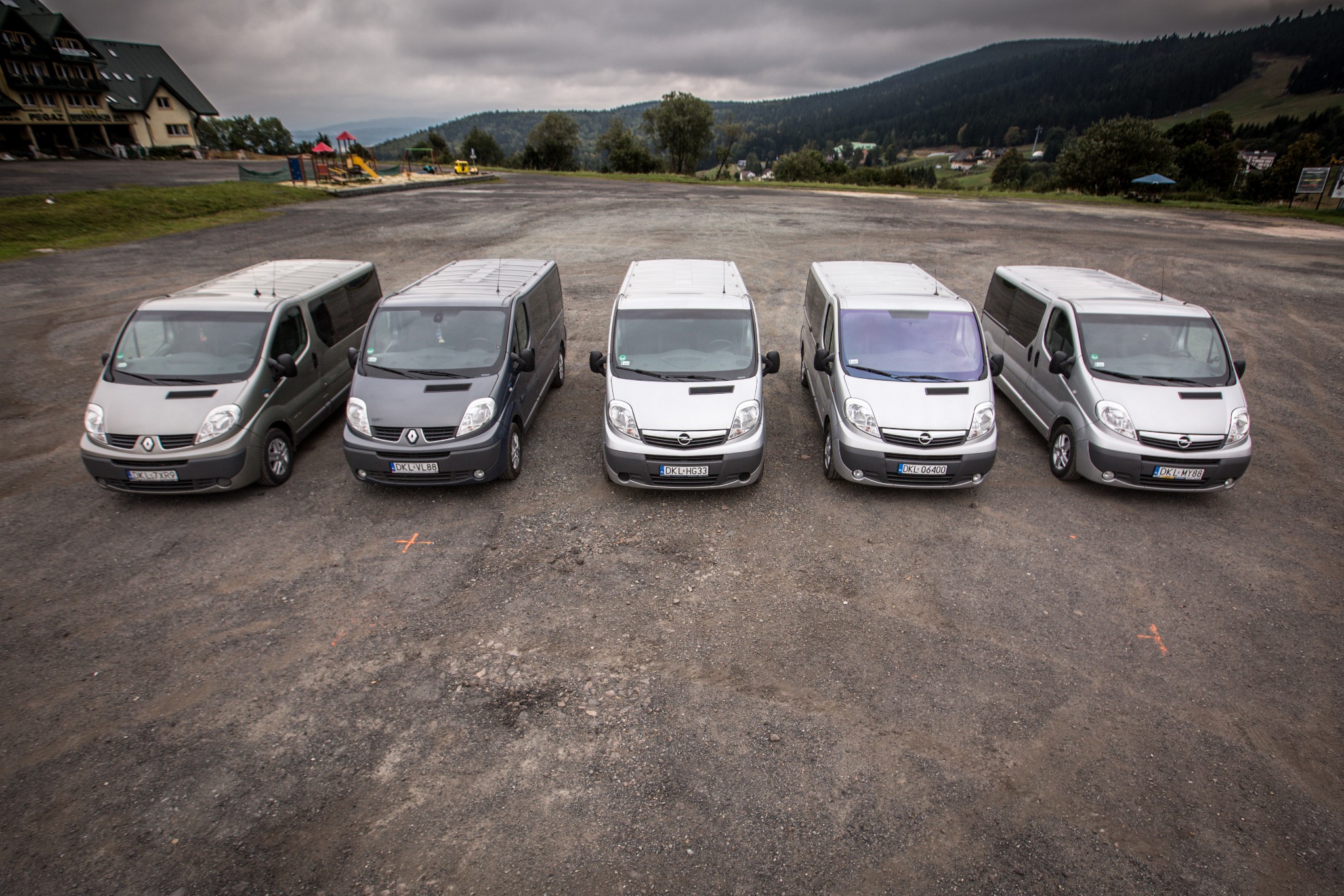 Rząd pięciu vanów różnych marek i kolorów (Renault Trafic i Opel Vivaro) zaparkowanych na żwirowym parkingu z budynkiem w tle i zalesionymi wzgórzami na horyzoncie, sfotografowanych z góry.