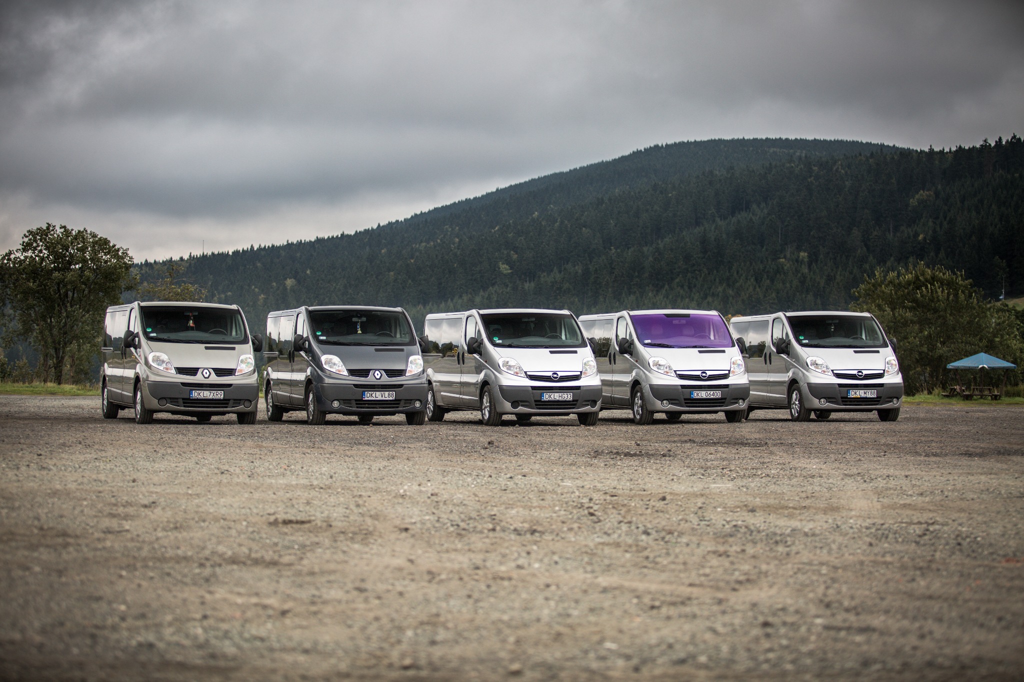 Rząd zaparkowanych busów różnych marek (Renault i Opel) na żwirowym parkingu z zalesionym wzgórzem w tle, jeden z busów ma fioletową szybę przednią.