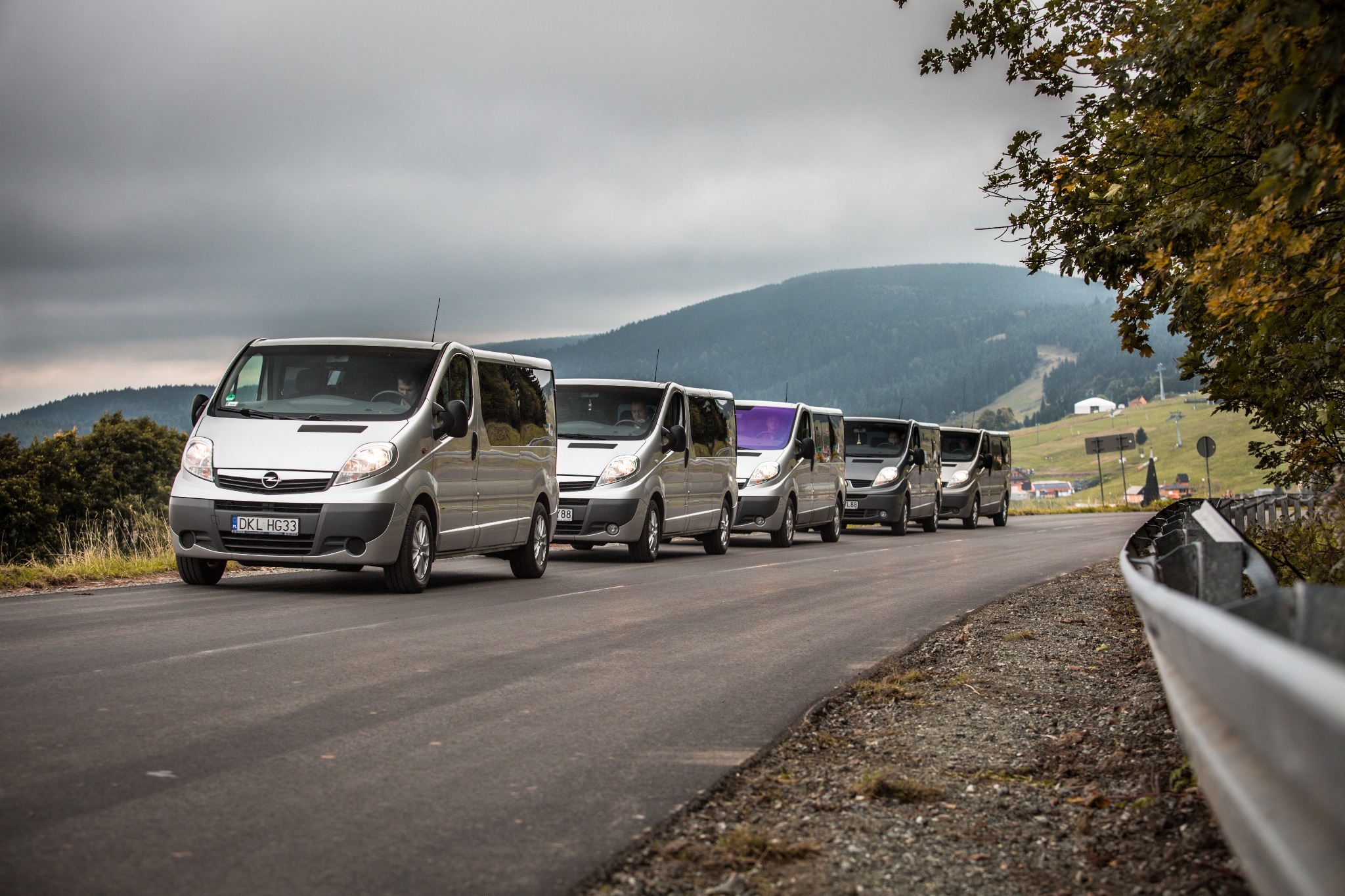 Kolumna srebrnych vanów Opel Vivaro jadąca po asfaltowej drodze na tle zalesionych wzgórz i pochmurnego nieba.