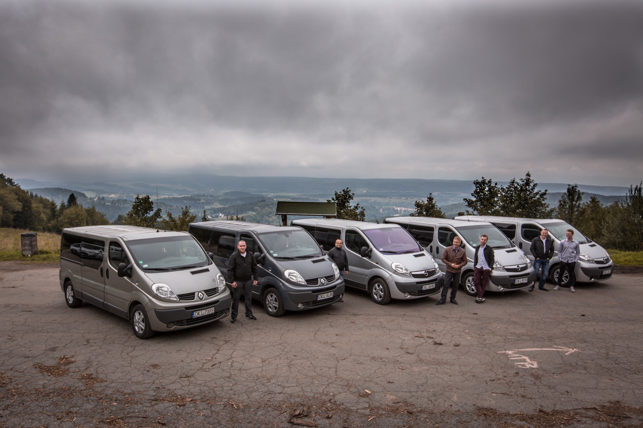 Flota srebrnych i szarych vanów Renault Trafic i Opel Vivaro z kierowcami na tle mglistego krajobrazu górskiego.