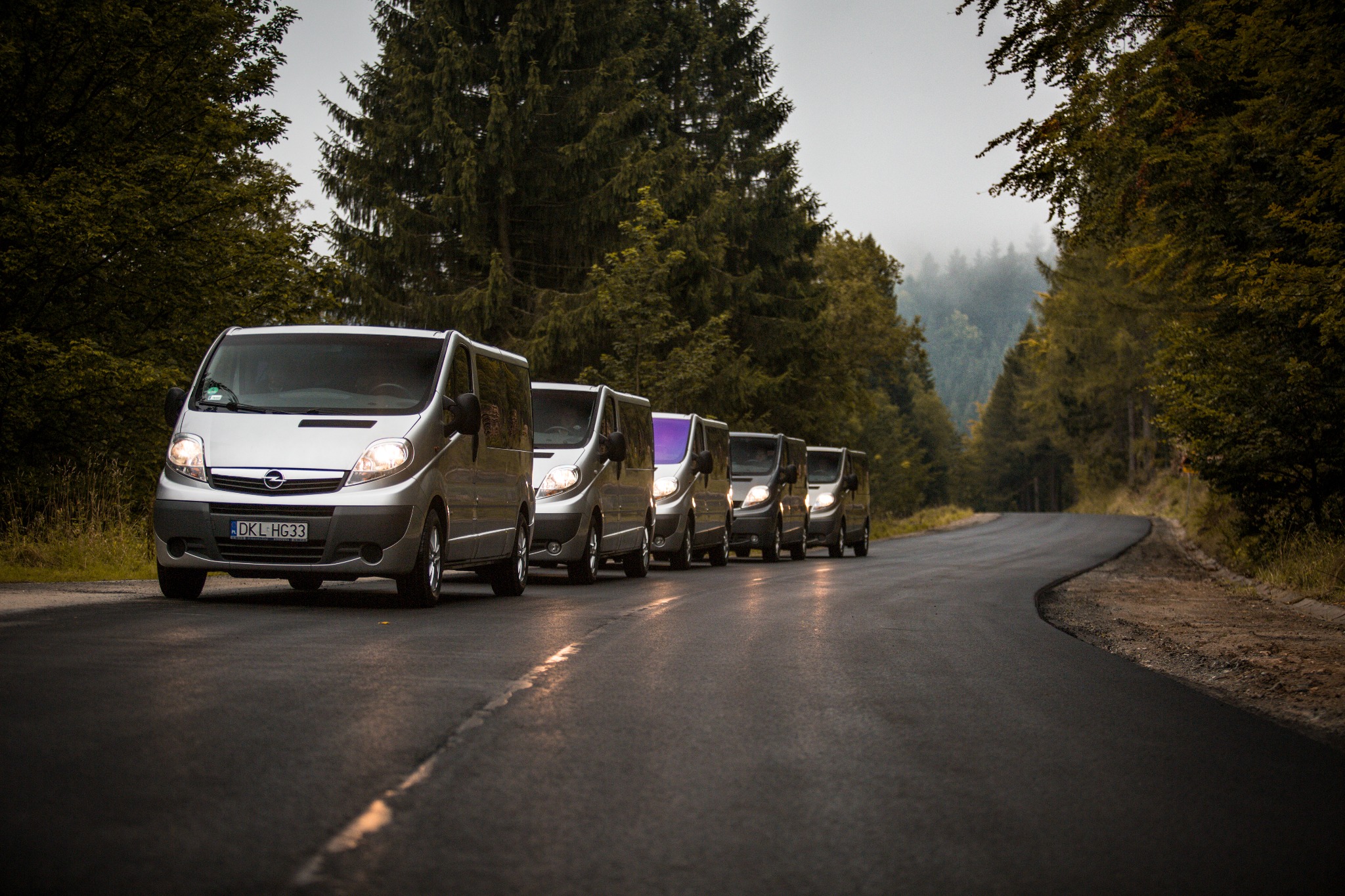 Szereg srebrnych vanów Opel Vivaro, jeden z fioletową szybą, jadących po mokrej, krętej drodze przez las w pochmurny dzień.