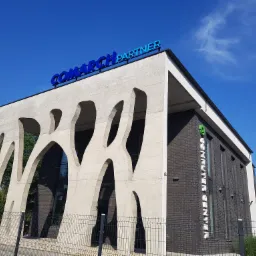 Budynek biurowy z napisem 'Comarch Partner' na dachu, betonowa fasada z abstrakcyjnymi wycięciami, ceglana ściana boczna z pionowym napisem, błękitne niebo.
