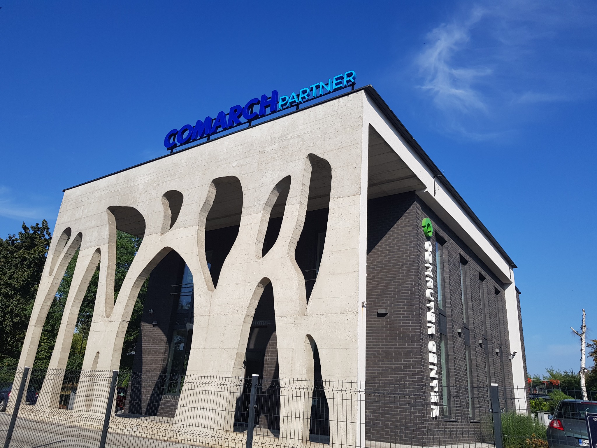 Budynek biurowy z napisem 'Comarch Partner' na dachu, betonowa fasada z abstrakcyjnymi wycięciami, ceglana ściana boczna z pionowym napisem, błękitne niebo.
